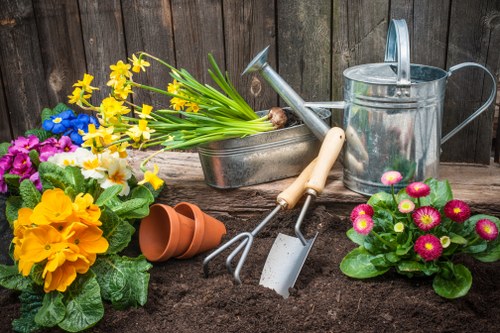 A neatly arranged garden scene features a freshly prepared flower bed with dark, rich soil in the foreground. To the left, vibrant flowering plants with bright yellow, pink, and purple blooms are in bloom, paired with lush green foliage. Behind the plants, a wooden fence provides a natural backdrop, with gardening tools including a watering can, a pair of gardening forks, a trowel, and a small garden shovel resting against it. Two empty terracotta plant pots are stacked beside a metal tray filled with yellow daffodil stems. The scene suggests an ongoing gardening activity, possibly the planting of new flowers or preparing for a spring garden overhaul, typical of landscaping and lawn care services in Bayswater. The natural lighting indicates a clear day, highlighting the vivid colours and textures of the garden elements, supporting professional gardening and landscaping efforts. This outdoor setting showcases well-maintained garden features suitable for outdoor maintenance and gardening services offered by Lawn Mowing Bayswater in the W2 postal area.
